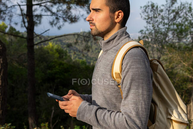 Вид сбоку на молодого бородатого этнического путешественника в повседневной одежде и рюкзаке на смартфоне, стоящего в пышном зеленом лесу во время похода в горной долине, отводя взгляд — стоковое фото