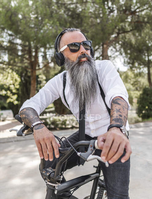 Confident male hipster in white shirt listening to music in headphones while sitting on bicycle on street with green trees — Stock Photo