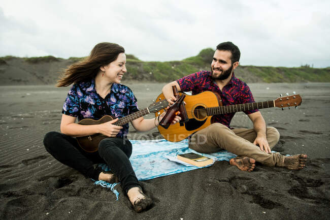 Positives Musikerpaar, das tagsüber mit Gitarren und klappernden Bierflaschen am Sandstrand am Meer sitzt — Stockfoto