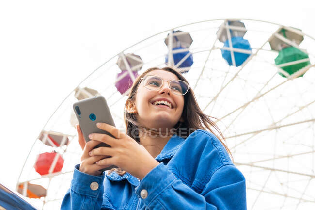 Улыбающаяся молодая женщина в повседневной одежде и очках, стоящая на городской улице во время сёрфинга по сотовому телефону рядом с металлическими перилами под колесами обозрения — стоковое фото