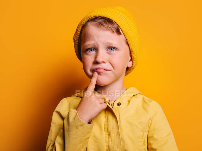 Thoughtful boy with blue eyes in vivid raincoat and beanie hat touching face and looking away against yellow background in studio — Stock Photo