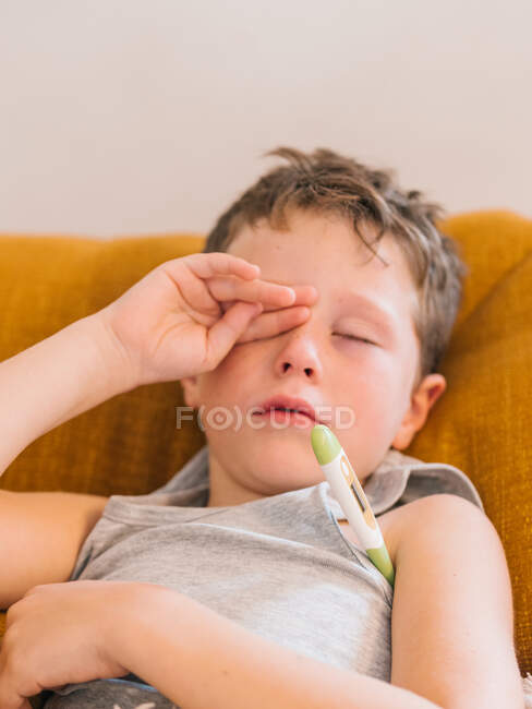 De cima de menino doente medir a temperatura com termômetro eletrônico enquanto deitado no sofá em casa e com gripe — Fotografia de Stock