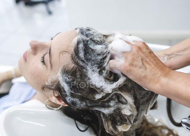 Cultivo irreconocible maestro lavado pelo oscuro de cliente femenino con los ojos cerrados en salón de belleza - foto de stock