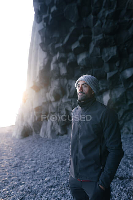 Задоволений молодий неголений етнічний чоловік подорожує в теплому одязі і капелюсі стоячи біля базальтових колон на пляжі Рейно з рукою в кишені і захоплюючись природою під час відпочинку в Ісландії. — стокове фото