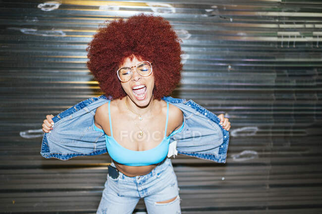 Delighted female with Afro hairstyle and stylish denim outfit with sunglasses standing near metal wall with closed eyes on dark street — Stock Photo