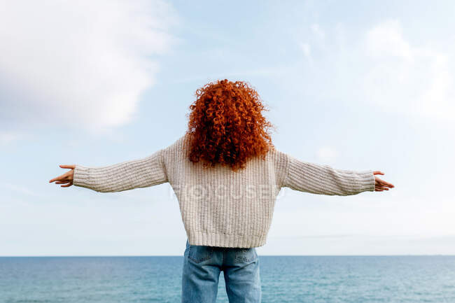 Обратный вид на неузнаваемую восторженную женщину с кудрявыми длинными рыжими волосами, поднимающими руки на берегу бурлящего моря — стоковое фото