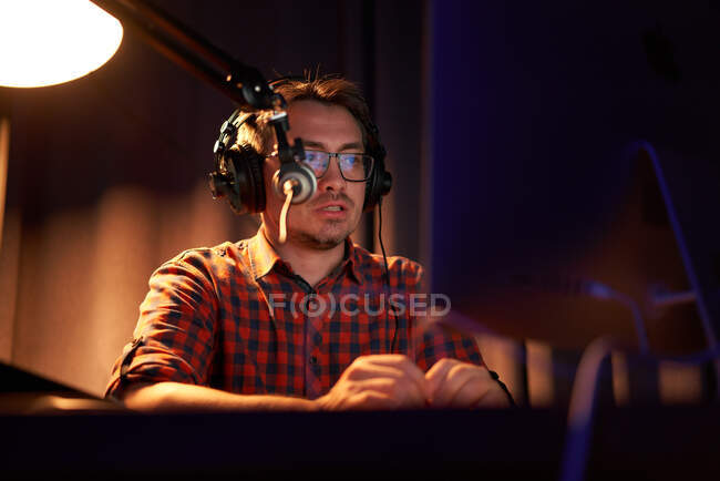 Desde abajo enfocado joven masculino en camisa a cuadros y anteojos usando computadora y hablando en micrófono mientras graba podcast en estudio oscuro - foto de stock