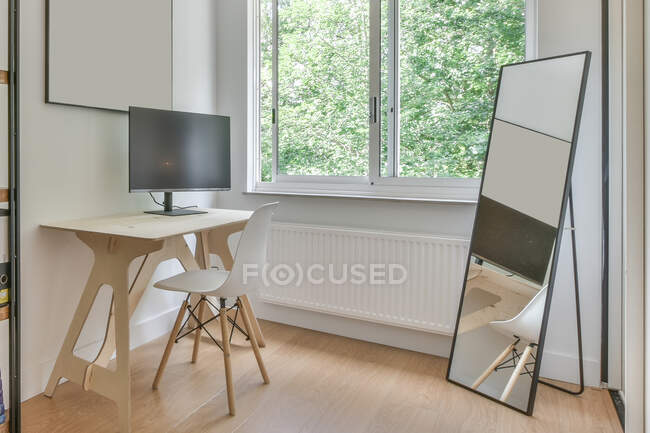 Стул за письменным столом с современным компьютером расположен возле окна с видом на деревья в светлой комнате с зеркалом с отражением в квартире — стоковое фото