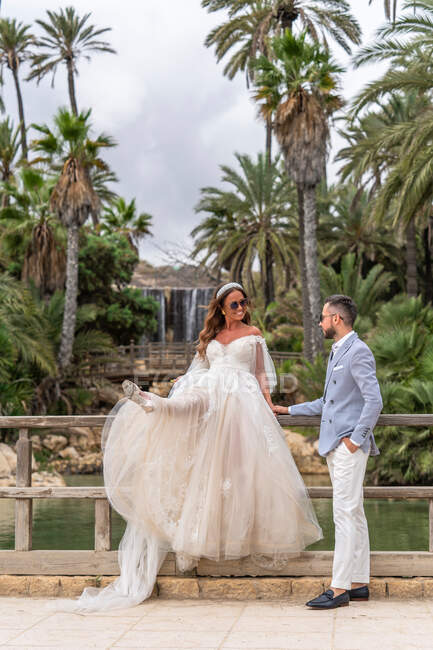 Jovem casal recém-casado em roupas de casamento em pé na passarela de madeira com trilhos perto do lago com pedras e palmas verdes e plantas no parque no dia de verão — Fotografia de Stock