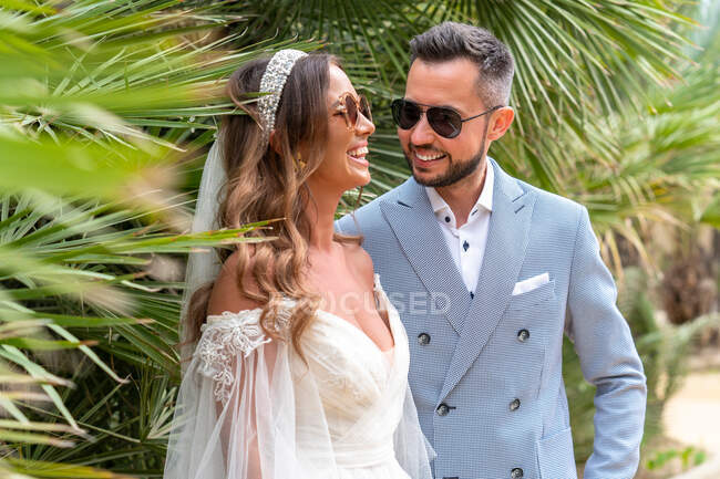 Sorridente giovane coppia sposata in abiti da sposa e occhiali da sole in piedi vicino a alberi verdi e piante in giorno d'estate nel parco — Foto stock