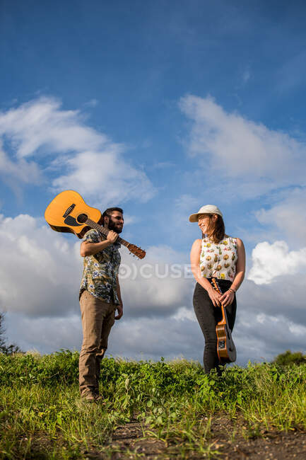 Низький кут веселого чоловіка музиканта з акустичною гітарою на плечі, що стоїть на зеленій траві біля жінки з укулеле в природі на тлі блакитного неба в сонячний день — стокове фото