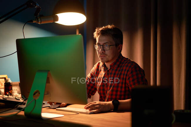 Homem concentrado em camisa quadriculada e óculos trabalhando no computador sentado à mesa com lâmpada e microfone durante a gravação podcast — Fotografia de Stock