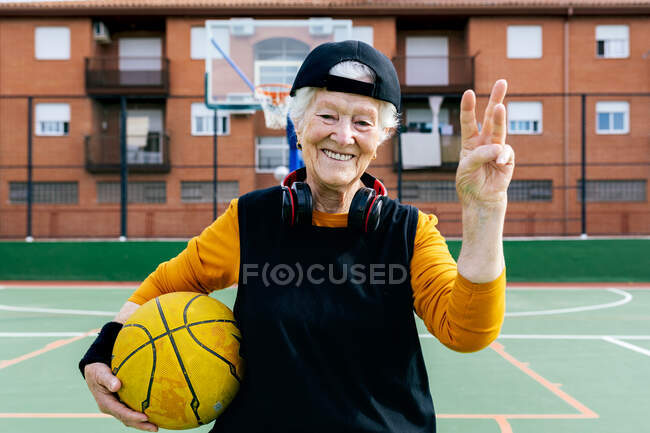 Mulher madura otimista em activewear e fones de ouvido olhando para a câmera enquanto está em pé na quadra de basquete pública com bola durante o treinamento — Fotografia de Stock
