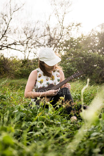 Mujer tranquila músico en traje casual y gorra sentado en la hierba verde y jugando ukelele en la naturaleza a la luz del día - foto de stock