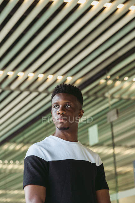Confiante afro-americano masculino em t-shirt casual de pé no edifício moderno e olhando para longe — Fotografia de Stock