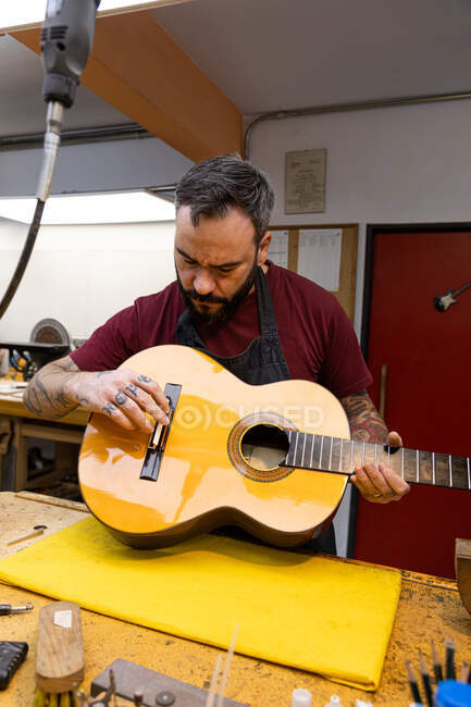 Мастер в фартуке настраивает гитару за столом с разными инструментами в ярком гараже — стоковое фото