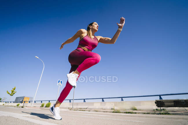 Angolo basso di femmina che corre veloce su strada asfaltata durante l'allenamento sotto il cielo blu — Foto stock