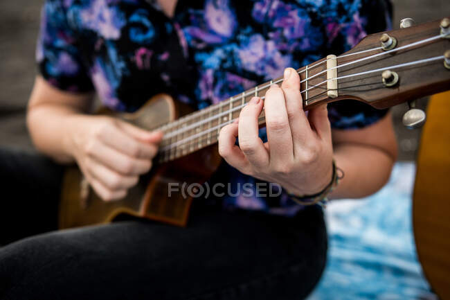 Cropped unrecognizable talented female musician with brown hair in casual clothes playing ukulele and singing song while sitting on sandy beach in nature in daylight — Stock Photo