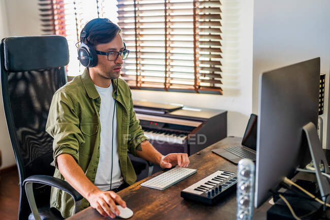 Vista lateral do cara concentrado com fones de ouvido sentado à mesa e usando o mouse enquanto trabalhava no computador — Fotografia de Stock