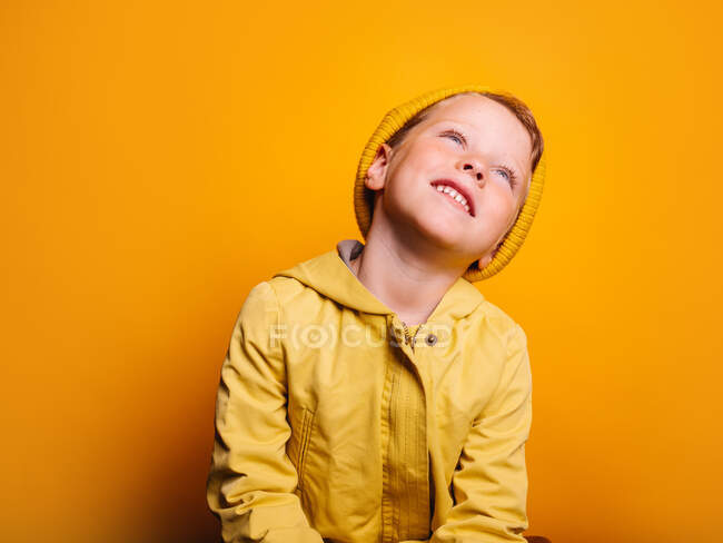 Happy boy in vivid yellow raincoat jacket and beanie hat laughing and looking away against yellow background in studio — Stock Photo