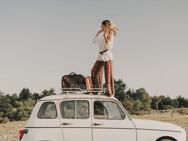 Full body side view of stylish female hippie in boho clothes standing on white old timer automobile with luggage during trip in nature — Stock Photo