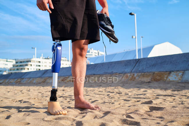 Uomo irriconoscibile con protesi alle gambe in abiti casual in piedi con le scarpe da ginnastica in mano sulla spiaggia di sabbia nella giornata di sole — Foto stock