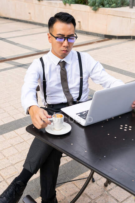 Wistful young Asian male entrepreneur with cup of hot drink and netbook looking at screen in urban cafeteria table in daylight — Stock Photo
