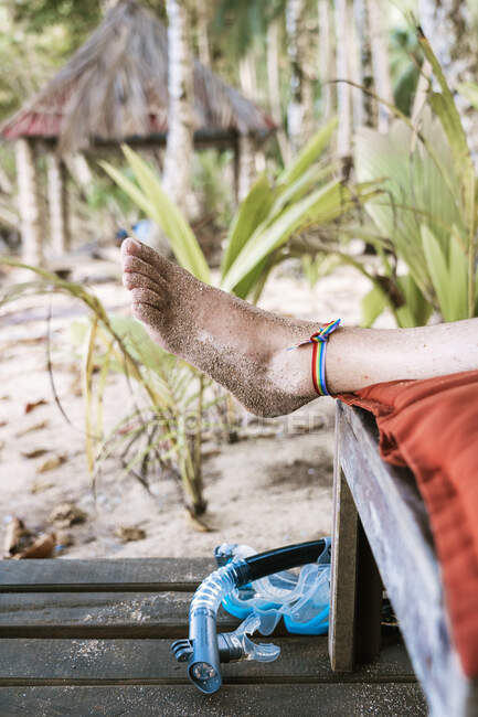 Bein mit Sand und Regenbogenband der Ernte unkenntlich Männchen liegt auf Bank vor tropisch grünem Hintergrund während Urlaub auf afrikanischer Insel — Stockfoto