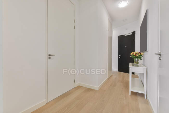 Pasillo con puerta blanca cerrada y jarrón de cristal con ramo de flores en luminoso apartamento moderno con puerta de entrada negra - foto de stock