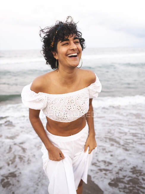 Позитивна етнічна жінка в білому стильному вбранні і з кучерявим волоссям, що стоїть на березі моря і сміється з закритими очима — стокове фото