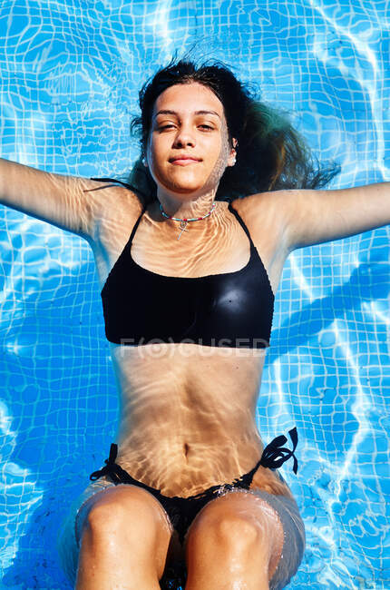 Von oben bezaubernde junge Frau im Bikini, die im Pool mit klarem Wasser schwimmt, während sie im Sommer in die Kamera schaut — Stockfoto