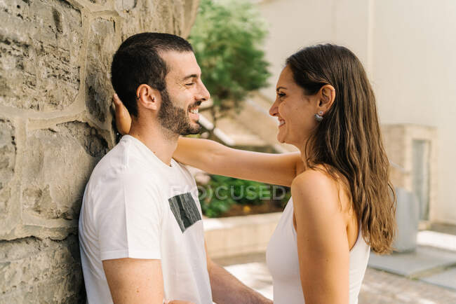 Side view of positive young ethnic woman in casual clothes embracing smiling bearded boyfriend leaning on aged stone building in city park on sunny day — Stock Photo