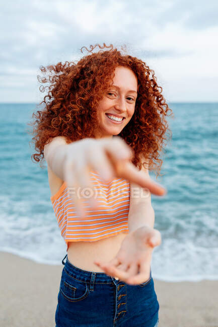 Piacevole femmina con lunga acconciatura riccia allo zenzero che raggiunge mano alla macchina fotografica mentre in piedi contro il mare ondulante — Foto stock