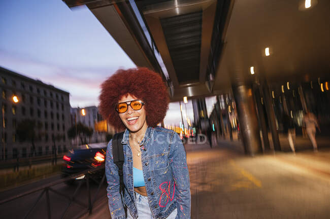 Positive Frau mit Afro-Frisur trägt stylisches Outfit mit Sonnenbrille und schlendert abends durch die Straßen mit Gebäuden in der Stadt — Stockfoto