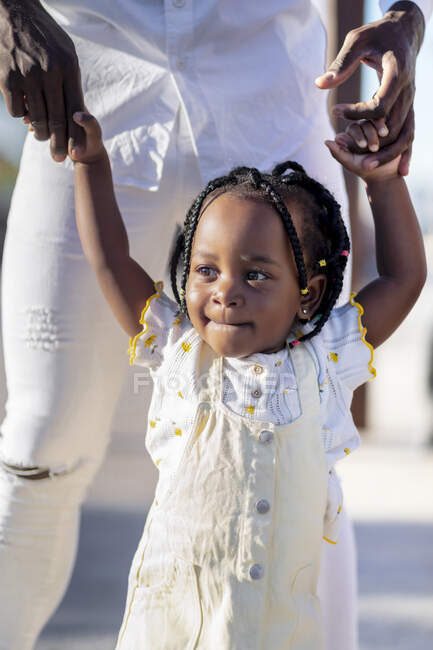 Happy African American little girl in light dress holding hands of unrecognizable father while walking on street in sunny day — Stock Photo