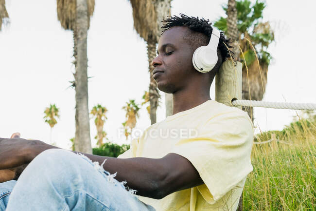 Afroamericano giovane maschio in abito casual ascoltare musica con le cuffie in giornata di sole nel parco con gli occhi chiusi — Foto stock