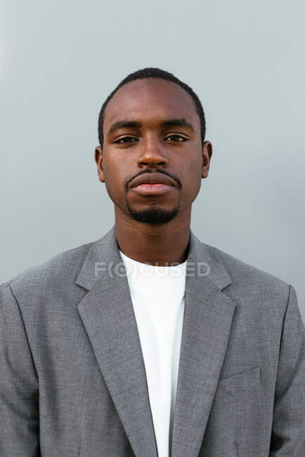 Empresario masculino afroamericano serio en traje formal de pie sobre fondo gris y mirando a la cámara - foto de stock