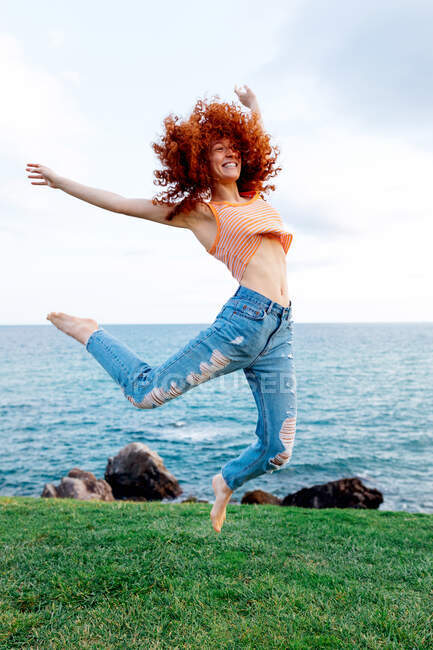 Cuerpo completo de hembra enérgica feliz con pelo rizado jengibre volador saltando por encima del suelo herboso en la costa de mar ondulado azul mirando hacia otro lado - foto de stock