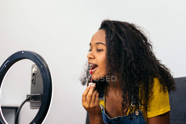 Mujer afroamericana aplicando lápiz labial mientras mira la cámara en el teléfono celular colocado en la luz del anillo - foto de stock