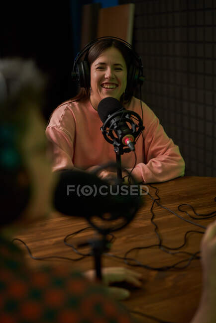 Joven presentadora de radio en ropa casual y auriculares sentada en la mesa con micrófono y comunicándose con su colega anónima durante la grabación de podcast en el estudio - foto de stock