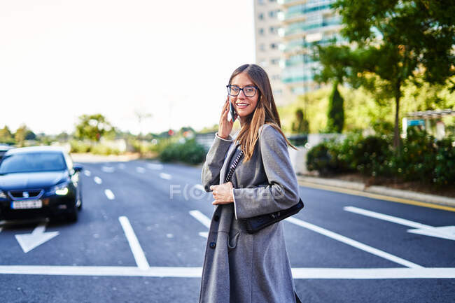 Vista lateral de la mujer de negocios positiva con abrigo de pie en la carretera de asfalto y tener llamada telefónica en el teléfono celular - foto de stock