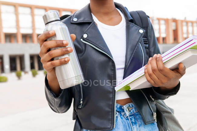 Анонимная афроамериканская студентка с термосом и кучей учебников, стоящих на улице возле здания университета — стоковое фото