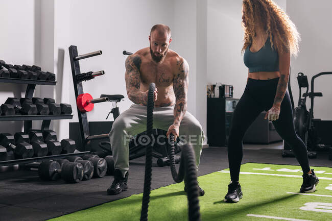 Мускулистый спортсмен с татуировками делает упражнения с веревкой во время функциональной тренировки с инструктором в спортзале в дневное время — стоковое фото