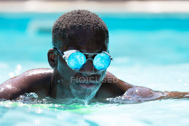 Спокойный афроамериканец в стильных солнцезащитных очках с отражением водного плавания в бассейне на размытом фоне в солнечный летний день — стоковое фото