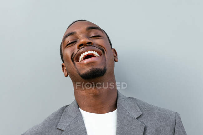 Feliz empresario afroamericano en traje formal riendo mientras está parado sobre fondo gris - foto de stock