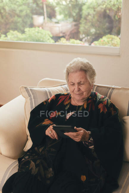 Focused senior female with gray hair resting on couch and reading e book on tablet in living room at home — Stock Photo