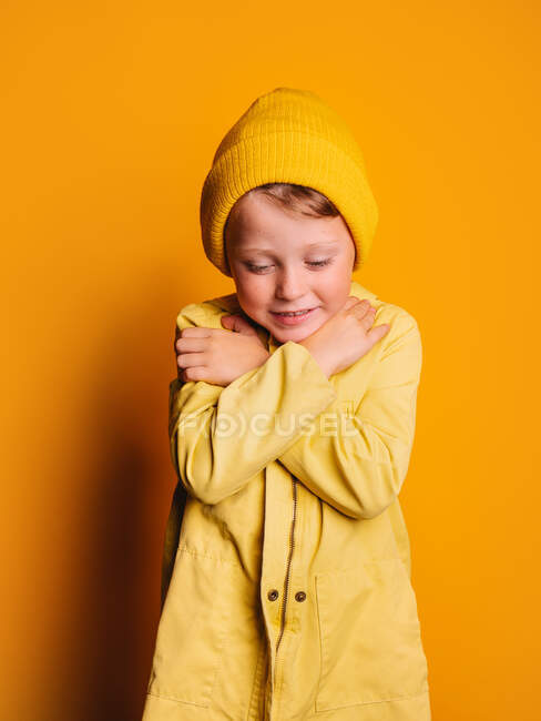 Agradable niño en impermeable de moda y gorro sombrero de pie con los brazos cruzados y mirando hacia abajo contra el fondo amarillo en el estudio - foto de stock