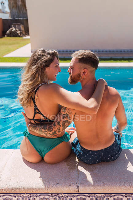 Back view of loving couple in swimsuits looking at each other while caressing on edge of swimming pool in tropical resort — Stock Photo