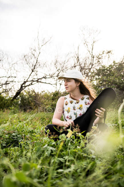 Спокійна жінка-музикантка в повсякденному одязі сидить на зеленій траві і відкриває чорний випадок акустичного укулеле в денне світло — стокове фото