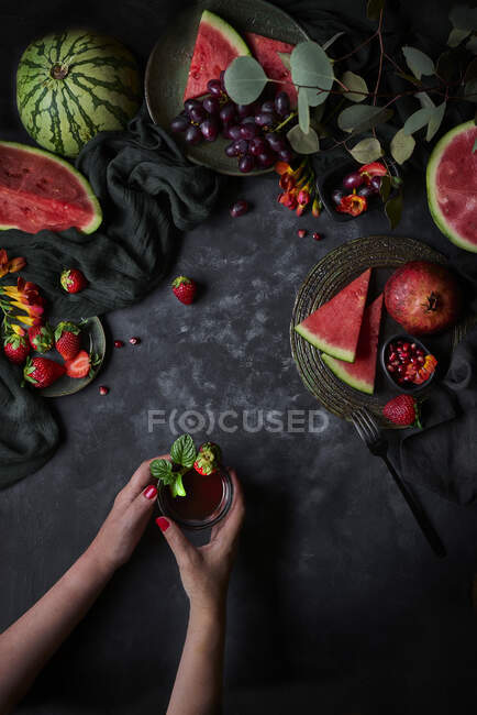 Top view of crop anonymous female with glass of beverage topped with strawberry and basil leaves on black background with watermelon and pomegranate — Stock Photo